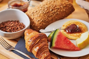 A breakfast setting with a croissant, bread, a bowl of cereal, a plate with a slice of watermelon, and a pancake with jam. - Les Trois Voiles (Port-Saint-Louis-du-Rhône)