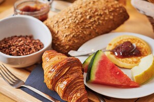 Disposição de café da manhã com um croissant, pão, uma tigela de cereal, um prato com uma fatia de melancia e um panqueca com geleia.