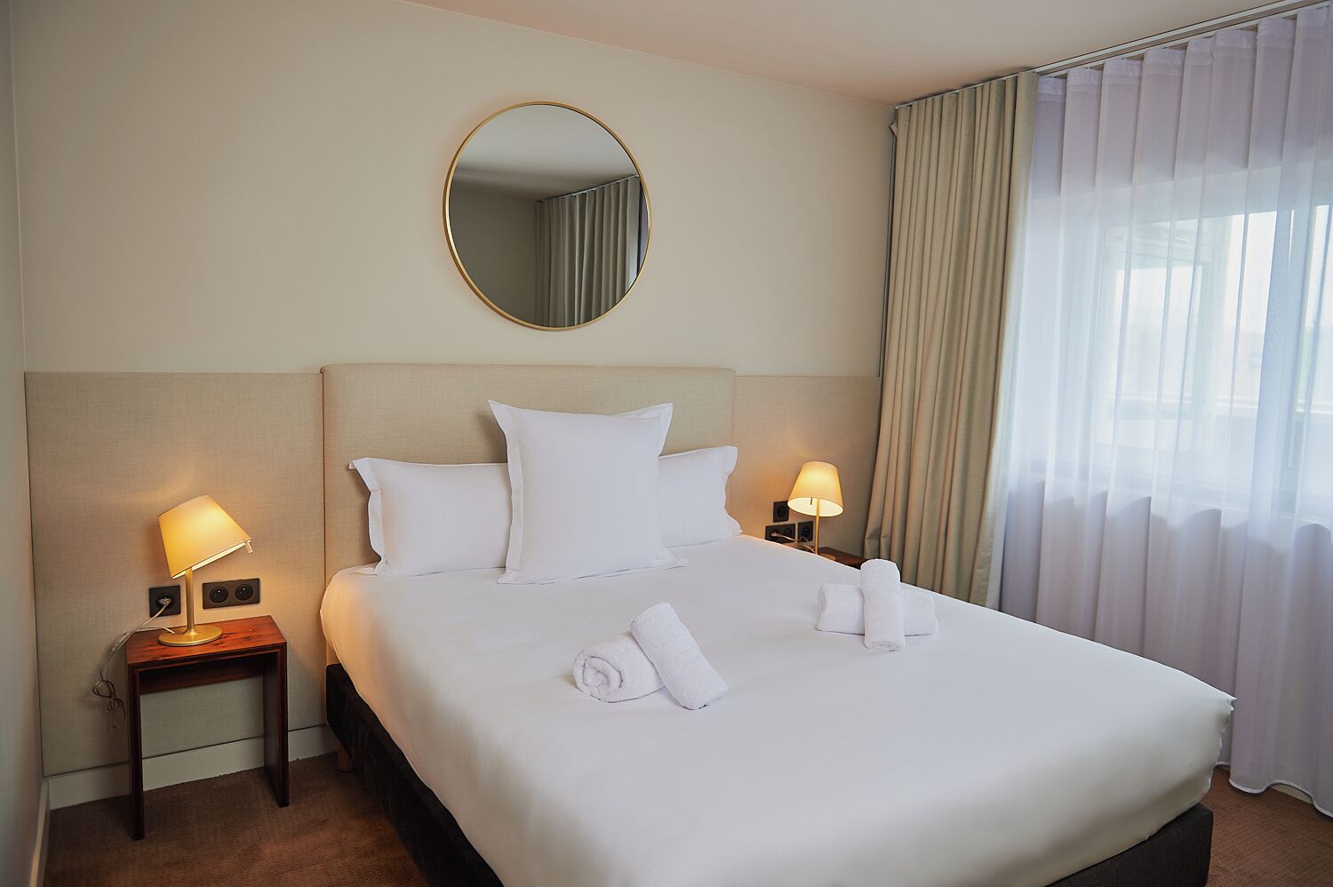 A hotel room with a bed, two bedside lamps, a round mirror, and a window with curtains.