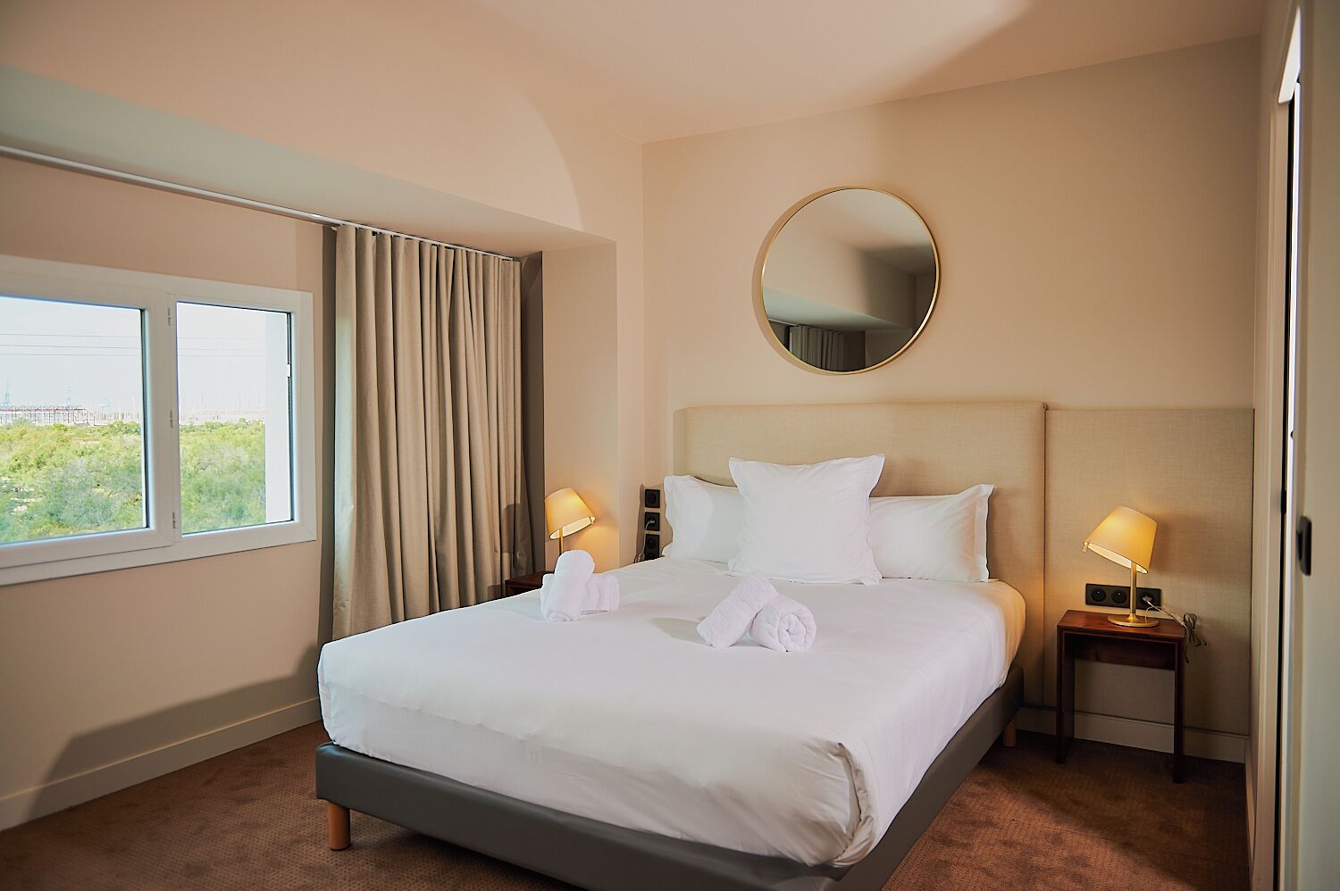 Ein Hotelzimmer mit einem großen Bett, zwei Nachttischlampen, einem runden Spiegel über dem Bett und einem Fenster mit Vorhängen.