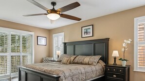 Ein Schlafzimmer mit einem dunklen Holzbett, einem Deckenventilator, einem Nachttisch mit Lampe und einem Fenster mit Jalousien.
