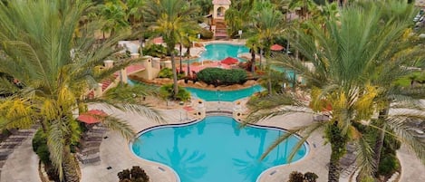 A large, kidney-shaped pool surrounded by palm trees and lounge chairs, with a small building in the background.