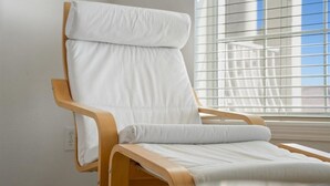A modern, wooden-framed lounge chair with white cushions, positioned near a window with blinds. - Villas Of Ocean Gate II with Ocean View (St. Augustine)