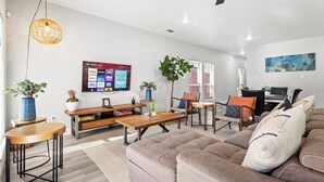 A modern living room with a sofa, coffee table, and TV. There are plants, a bookshelf, and a painting on the wall. - Modern home in Grand Prairie near DFW (Grand Prairie)