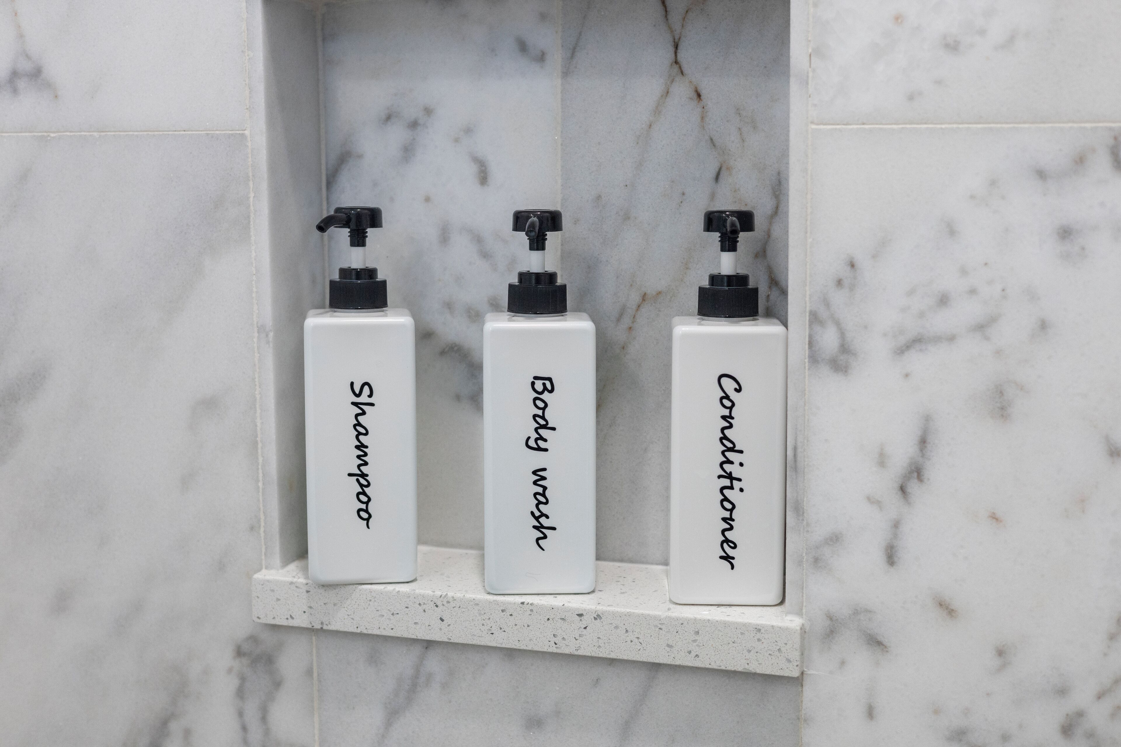 Three bottles of shampoo, body wash, and conditioner on a marble shelf.