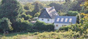 Une maison avec un toit en pente, entourée d’une végétation luxuriante et d’arbres.