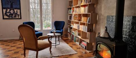 A cozy living room with a fireplace, a bookshelf, two chairs, a round table, and a large window.