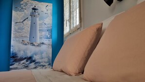 A room with a large painting of a lighthouse, a window with a view, and a beige sofa. - Grand Large (LE FAOUET)