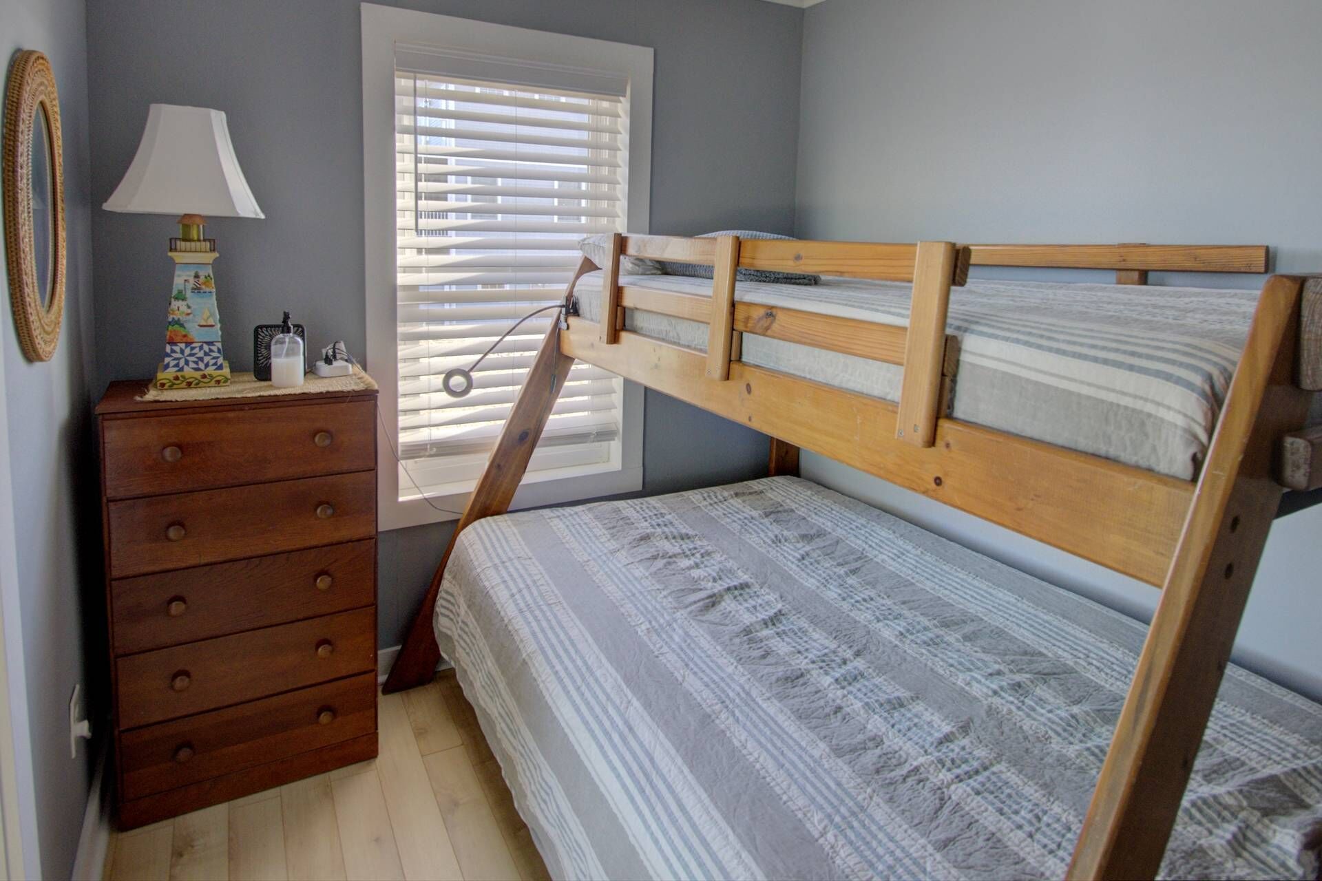 Une chambre avec un lit superposé, une commode en bois, une lampe et une fenêtre avec des stores.