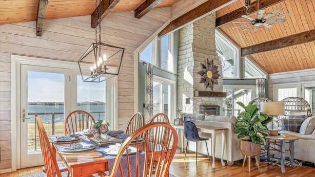 Un comedor amplio con sillas de madera y una mesa, ventanas grandes con vista a la playa y una chimenea de piedra.