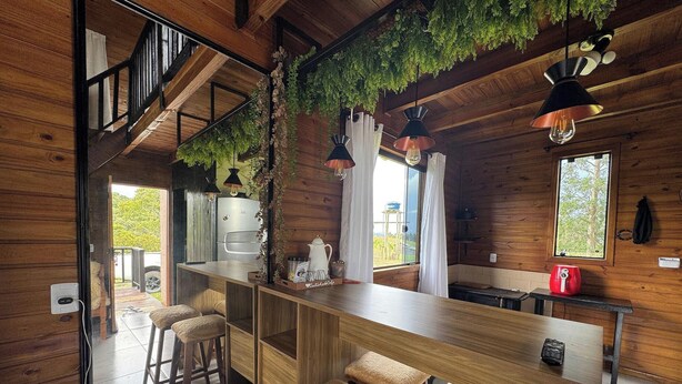 Interior d'una cabana de fusta amb cuina, tamborets de bar, plantes penjants i una escala.