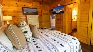 Ein Zimmer in einer Holzhütte mit einem Bett, einem Schminktisch, einem Fernseher und einer Tür zu einem weiteren Raum.