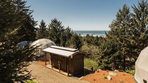 Un terrain de camping avec une petite structure en bois et une tente en forme de dôme, entouré d’une forêt dense et offrant une vue sur une étendue d’eau.