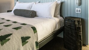 Un lit avec des oreillers blancs et à motifs, une couverture verte et blanche, une tête de lit bleu clair, un luminaire fixé au mur et une poubelle.