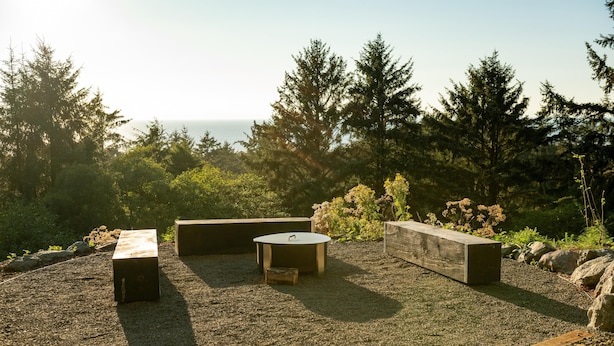 Un espace extérieur aménagé avec des bancs et une table ronde, entouré de gravier et de rochers, avec des arbres et une vue sur l’océan en arrière-plan.