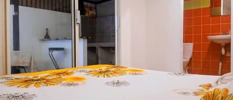 Un dormitorio con cama y colcha estampada, puerta corrediza de vidrio que da a un balcón, y baño con lavamanos e inodoro.
