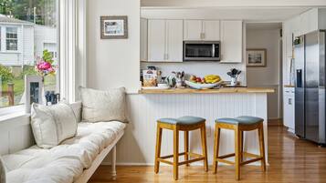 A kitchen with white cabinets, a microwave, and a refrigerator. There is a wooden bench with two stools, a window with a view of greenery, and a vase with flowers on the windowsill.
