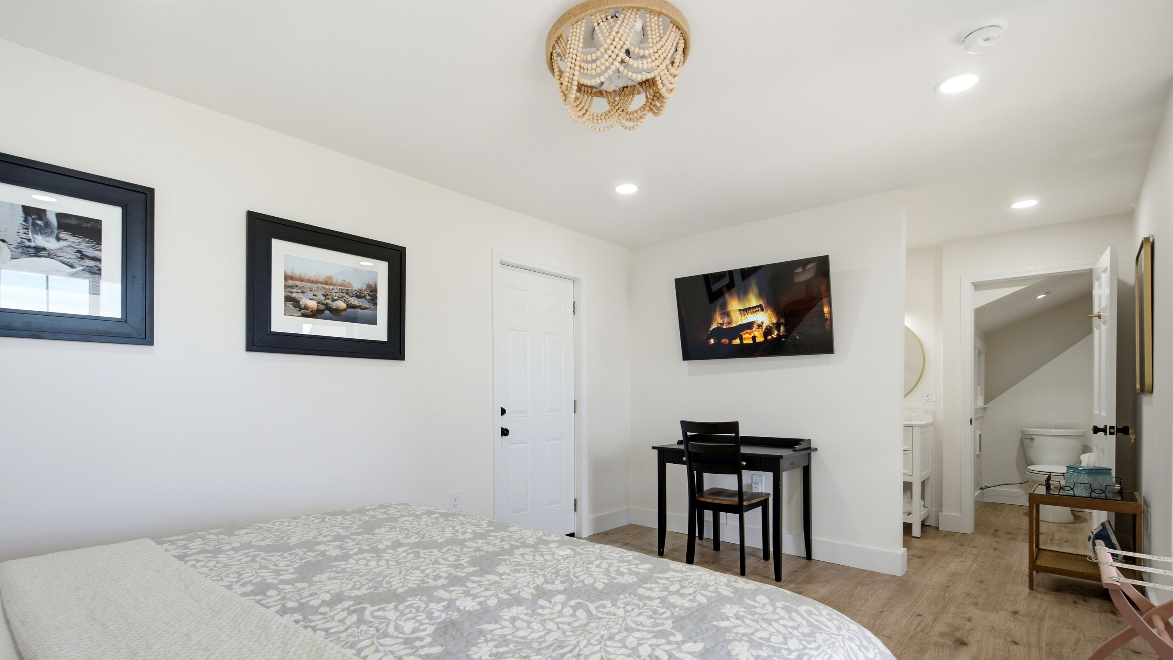 A bedroom with a bed, a desk, and a chair. There are framed pictures on the wall, a chandelier, and a painting of a fire.