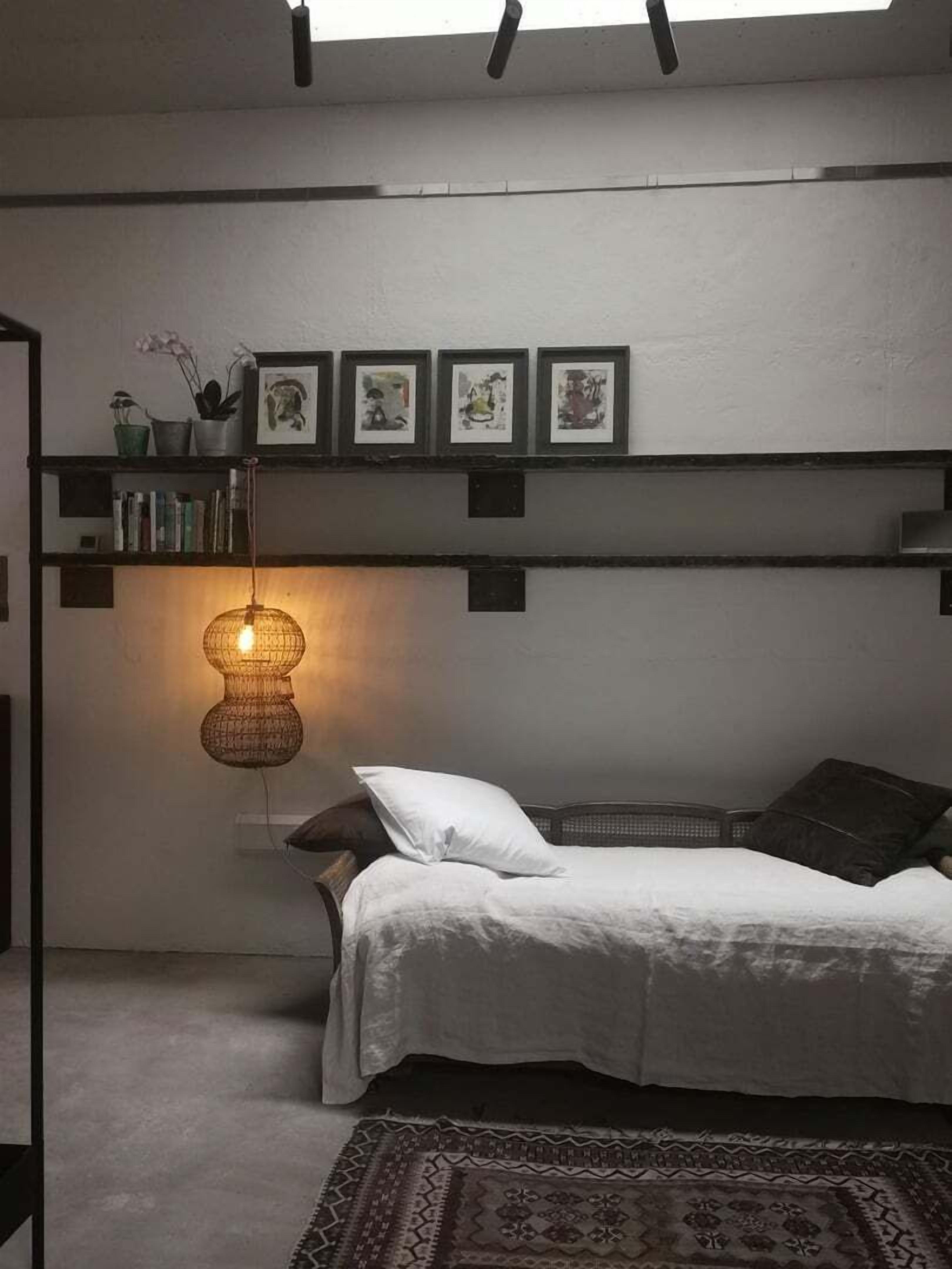 Une chambre à coucher avec un lit, une lampe suspendue, une étagère avec des photos encadrées et une fenêtre avec des stores.