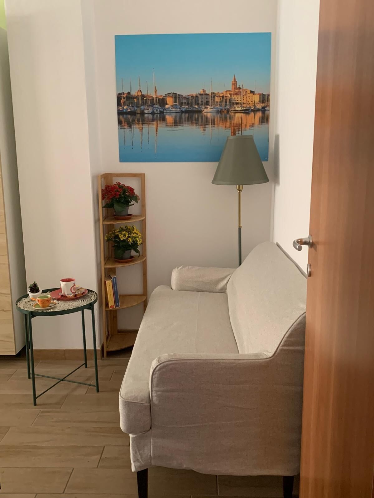 A room with a sofa, a small table with a lamp, a shelf with flowers, and a painting of a harbor.