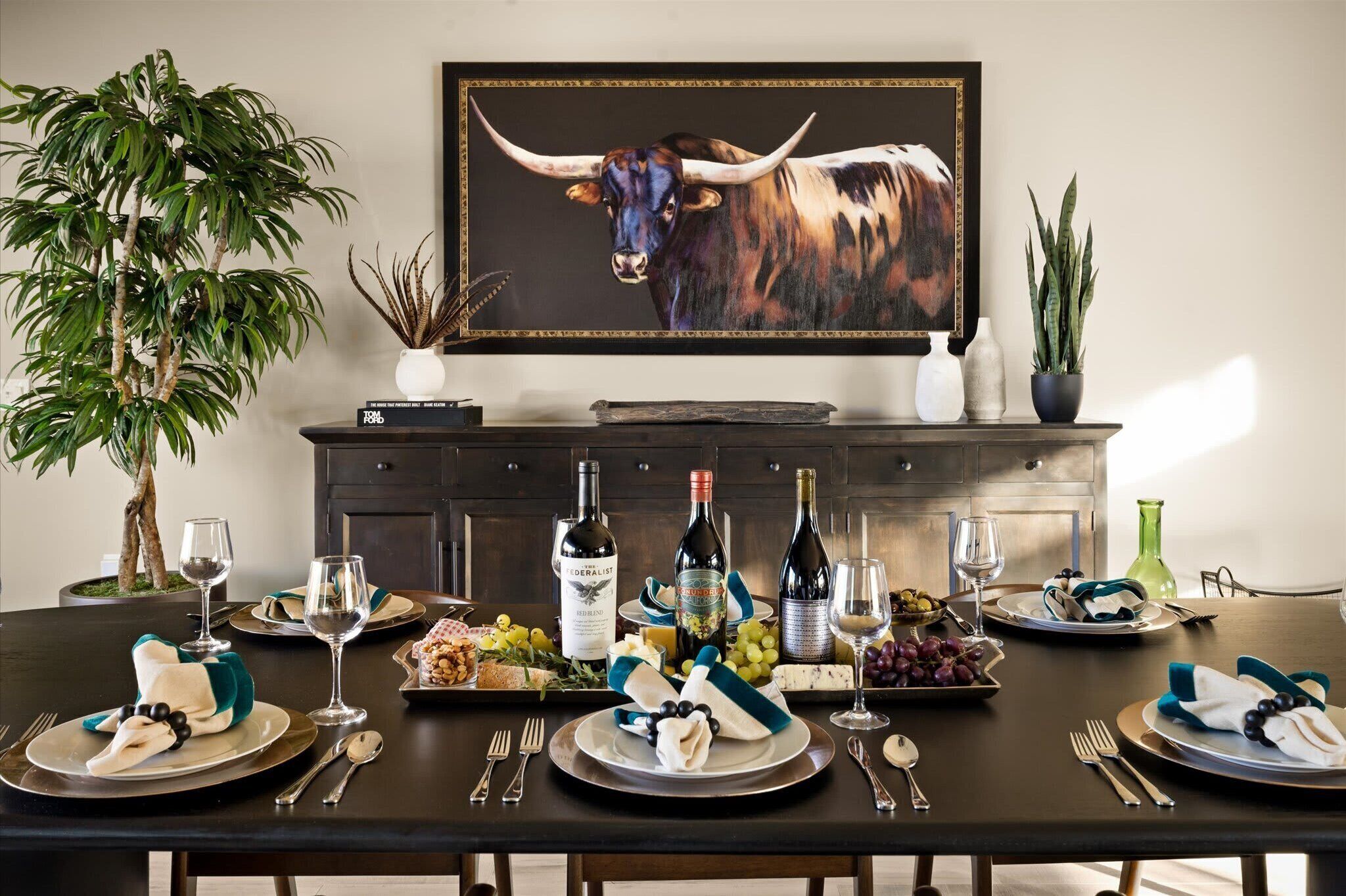 Une table à manger dressée avec des assiettes, des verres et des bouteilles, entourée de plantes en pot et d’une œuvre d’art encadrée.