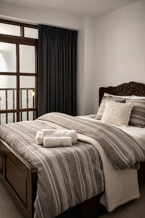 A neatly made bed with striped bedding and a wooden headboard, a window with a view, and a dark curtain.