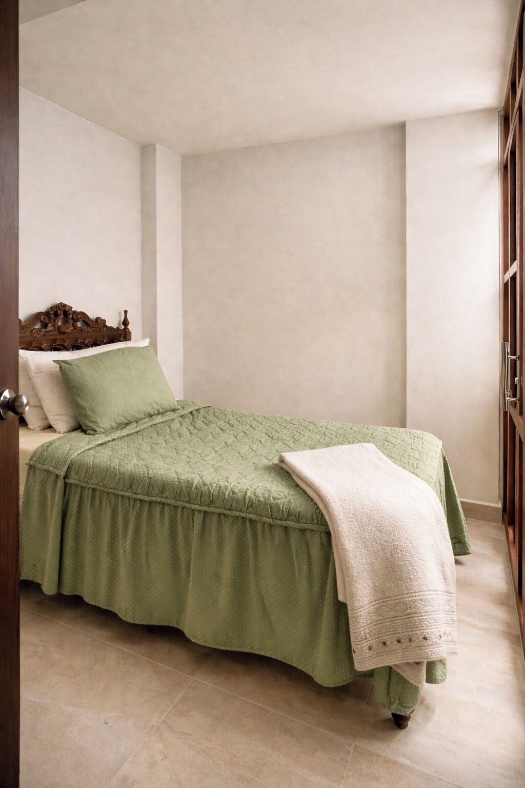A neatly made bed with a green bedspread and a white blanket, a wooden headboard, and a door to the left.