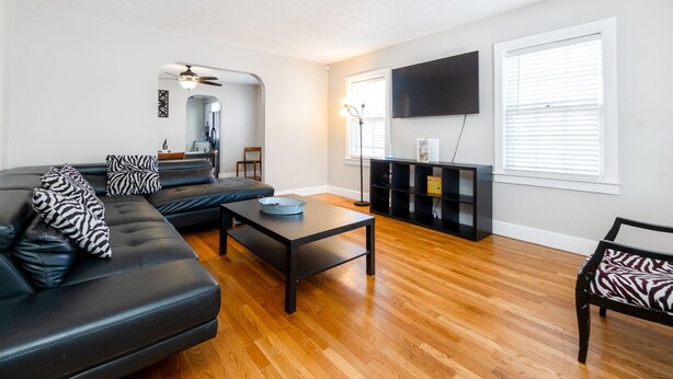 A modern living room with a black leather sofa, a coffee table, and a zebra-patterned bench. There is a flat-screen TV mounted on the wall and a black entertainment unit below it. - 5BR Home Near Airport, Expo, Downtown Sleeps 12 (Louisville)