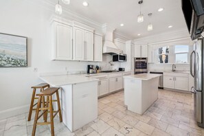 A modern kitchen with white cabinetry, a central island, bar stools, and stainless steel appliances. - Gulf Front Home with Private Pool, In-Home Elevator and Movie Theatre (Miramar Beach)