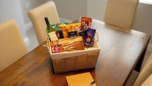 Ein Holztisch mit einem Korbwägelchen voller Snacks, einer Weinflasche und einer Schokoladentafel. Außerdem ist ein Notizblock mit einem Stift zu sehen.