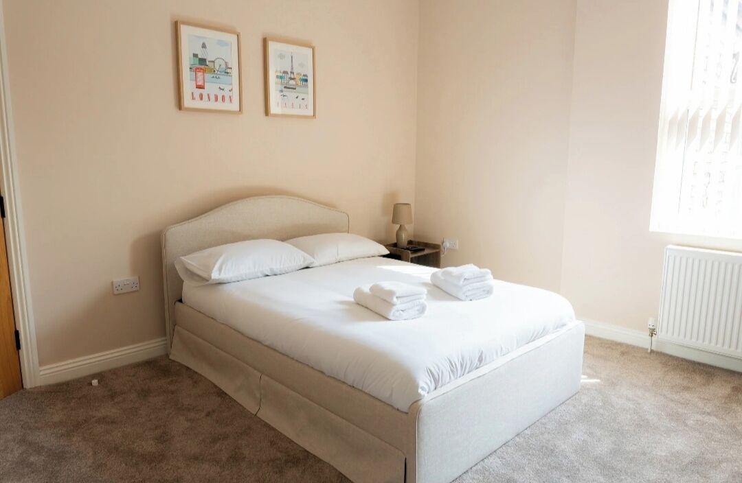 Een keurig opgemaakt bed met witte lakens en handdoeken, een nachtkastje met een lamp, en twee ingelijste foto's aan de muur.