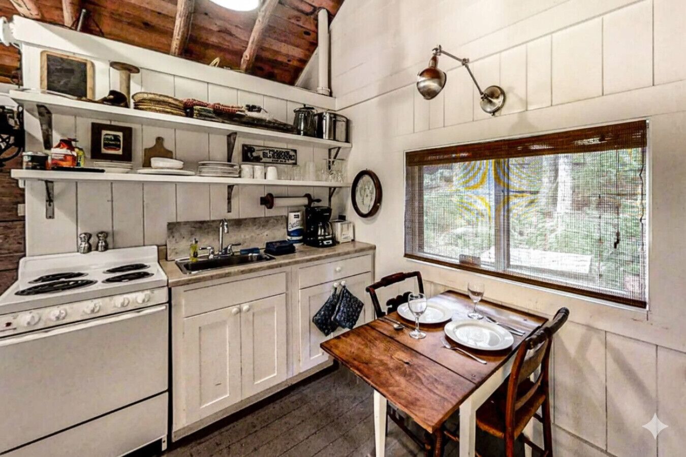 Una cocina con armarios blancos, estufa, cafetera y una mesa de comedor lista para dos personas.