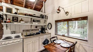 A kitchen with white cabinets, a stove, a coffee maker, and a dining table set for two. - Sprucewold Beach House | Romantic Coastal Escape (Boothbay Harbor)