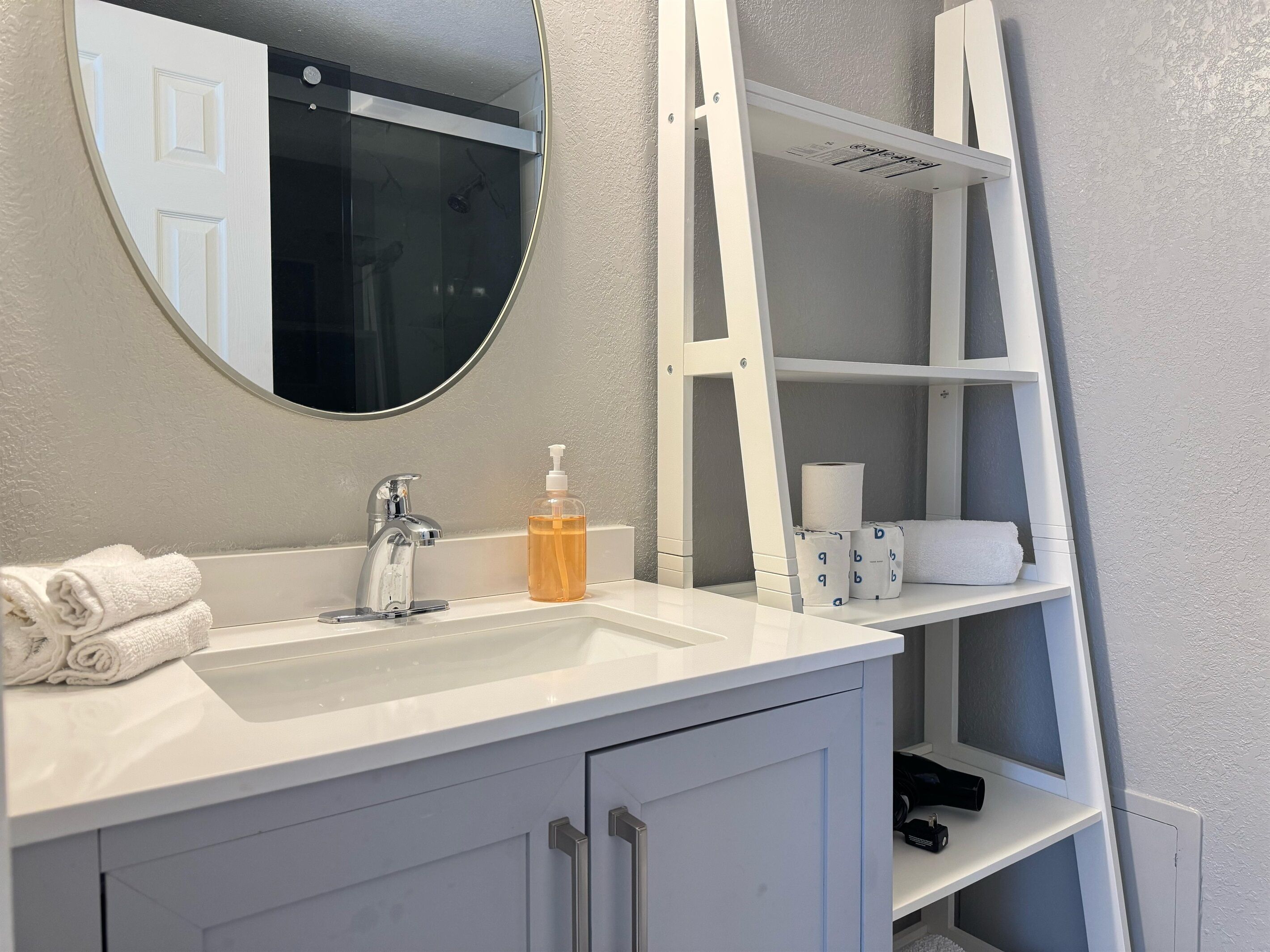 Uma casa de banho com toucador branco, um espelho redondo, uma prateleira branca com produtos de higiene e uma toalha branca no balcão.