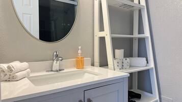 Uma casa de banho com toucador branco, um espelho redondo, uma prateleira branca com produtos de higiene e uma toalha branca no balcão.