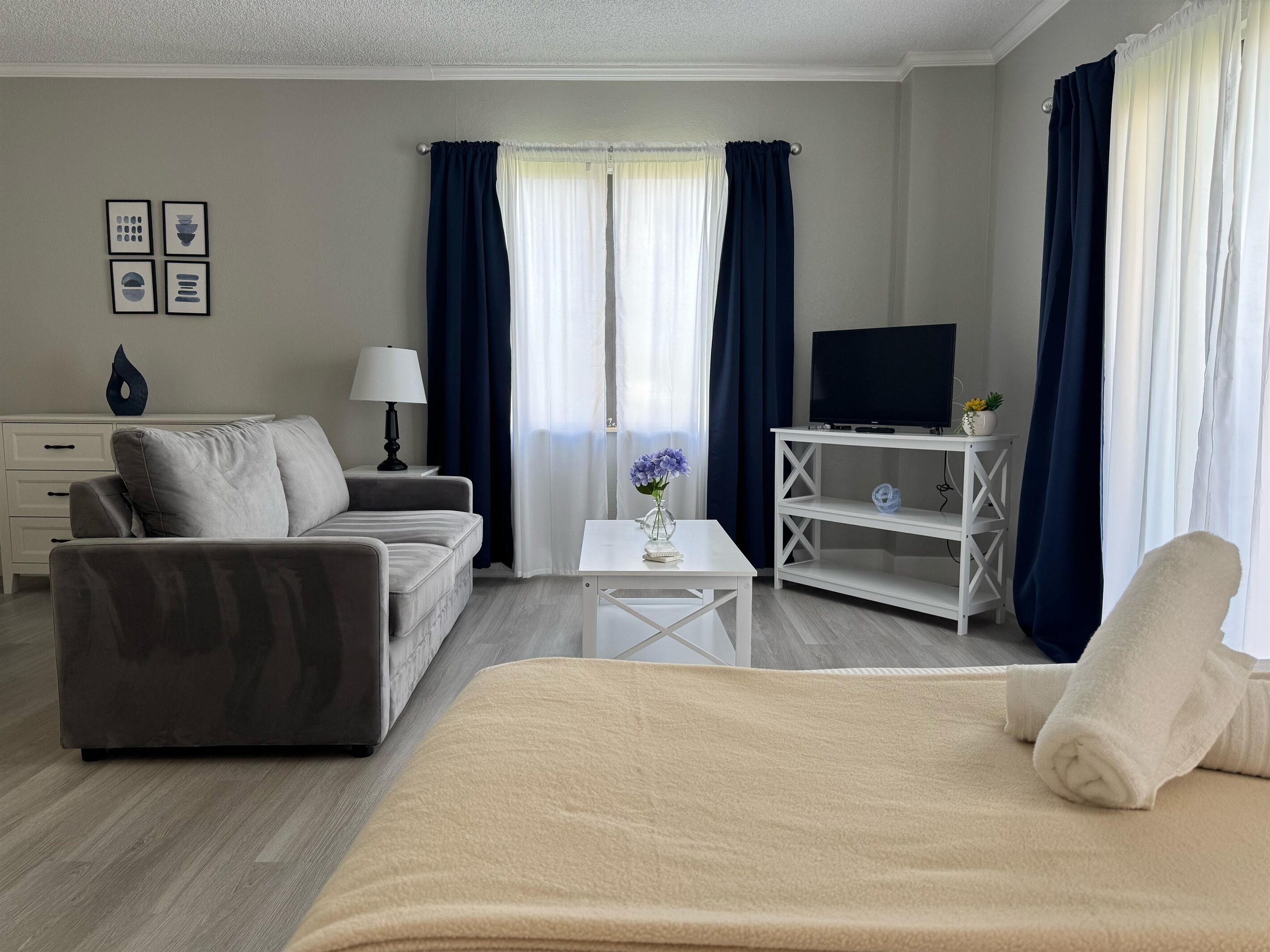 A modern hotel room with a bed, a sofa, a TV on a white stand, a small table with a vase, and a window with curtains.
