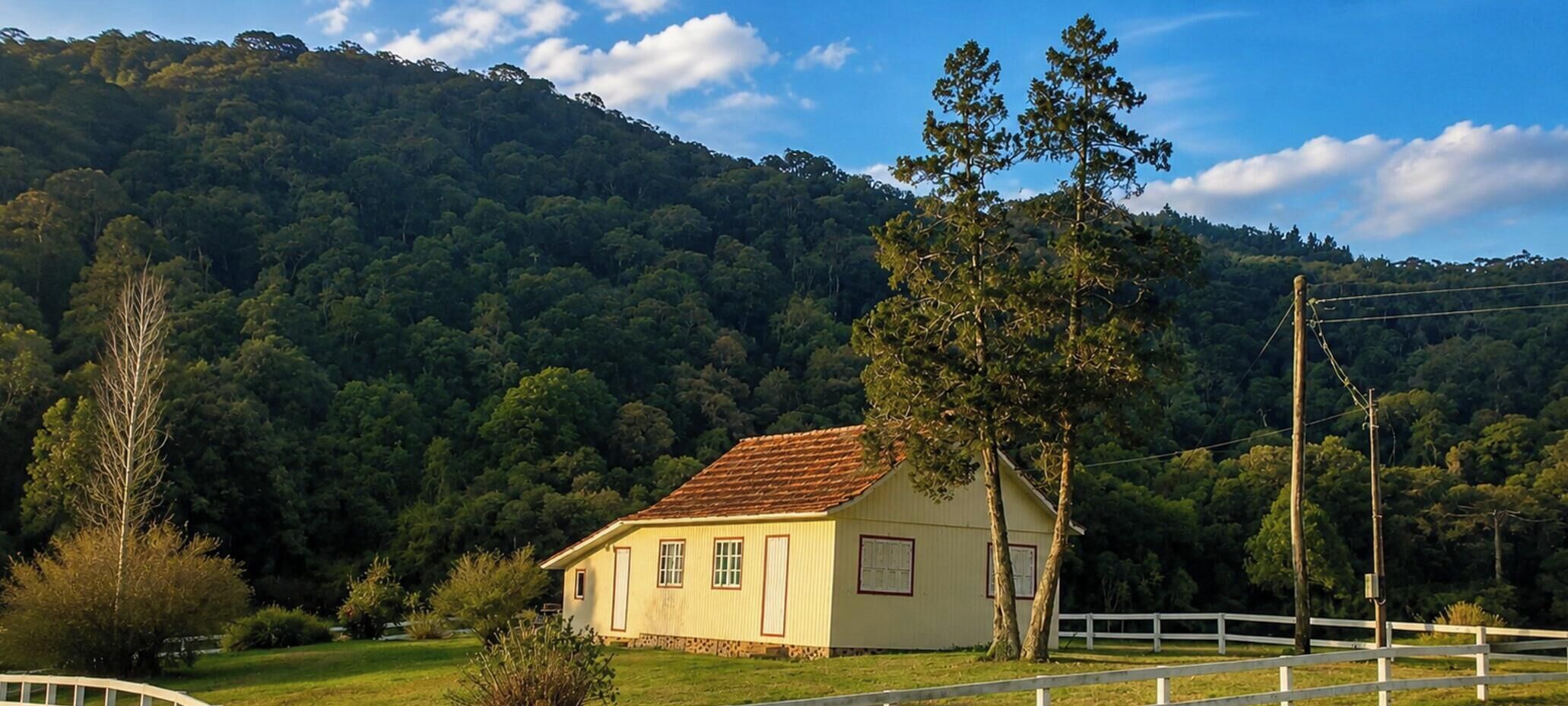 Uma casa térrea com telhado de telhas vermelhas, cercada por um muro branco, com uma colina arborizada ao fundo.