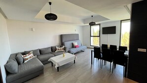 Ein modernes Wohnzimmer mit einer grauen Eckcouch, einem Couchtisch und einem Essbereich mit Tisch und Stühlen. Ein großes Fenster lässt viel Tageslicht herein und es ist ein Flachbildfernseher an der Wand montiert.