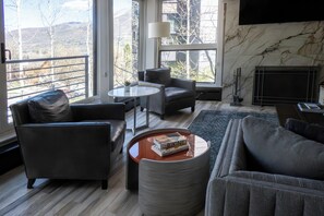 Ein modernes Wohnzimmer mit einer Couch, einem Sessel, einem Couchtisch, einem Kamin und einem großen Fenster mit Blick auf Bäume und Gebäude.