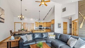 A spacious living room with a large L-shaped black leather sofa, wooden coffee table, and a ceiling fan. A kitchen with wooden cabinets and a dining area with a wooden table and chairs are visible in the background. - Flathead Lodge | Close to Skiing & Glacier Park (Somers)