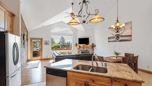 A modern kitchen with granite countertops, wooden cabinets, and a central island with a sink. There is a dining area with a table and chairs, and a large window offering a view of the outdoors. - Flathead Lodge | Close to Skiing & Glacier Park (Somers)