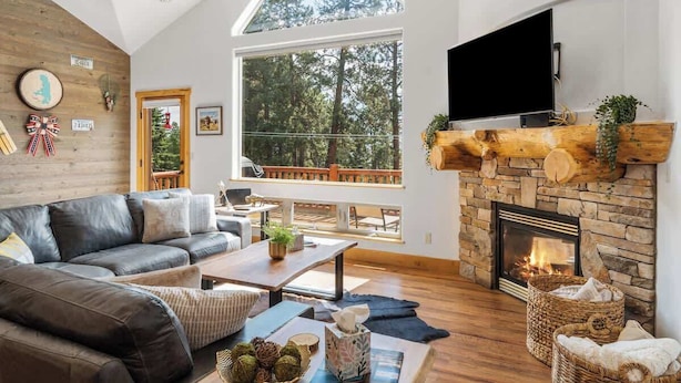 A cozy living room with a fireplace, a large window with a view of trees, a leather sofa, and a flat-screen TV mounted above the fireplace. - Flathead Lodge | Close to Skiing & Glacier Park (Somers)
