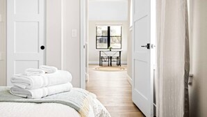 A hotel room with a bed, a stack of white towels, a wooden headboard, and a view of a dining area through an open door. - The Golden House | 1 Block to Main St (Golden)