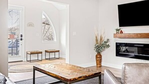 A modern living room with a herringbone-patterned coffee table, a leather sofa, a flat-screen TV mounted above a fireplace, and a large window with a view of the outside. - The Golden House | 1 Block to Main St (Golden)