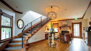 A spacious room with a wooden staircase, a round mirror, a dining area with a table and chairs, and a large window.