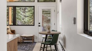 Un interior moderno con techo de madera, ventanas amplias que dan a un bosque, una zona de comedor con mesa y sillas, y un banco.