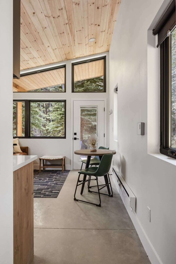 A modern interior with a wooden ceiling, large windows offering a forest view, a dining area with a table and chairs, and a bench.
