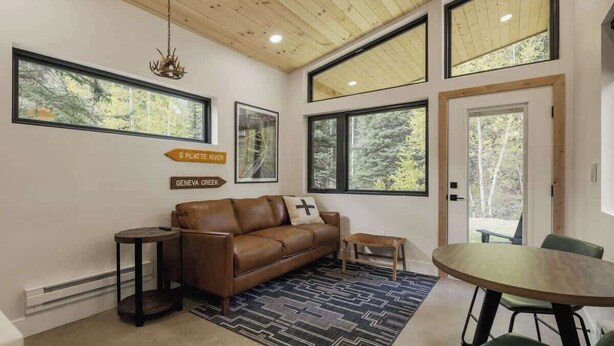 Una sala de estar moderna con un sofá de cuero marrón, una mesita de madera pequeña y una mesa de comedor con sillas. Hay ventanas amplias que ofrecen vistas a árboles y un letrero que indica 'Platte River' y 'Geneva Creek'.