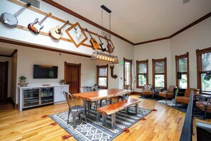 A spacious living room with a wooden dining table, chairs, and a large TV. There are multiple windows with blinds, a ceiling with hanging guitars, and a kitchen area with cabinets and a refrigerator. - Smiths Crescendo | Curtis Park Club | RiNo (Denver)