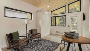A spacious room with a ceiling fan, large windows, a wooden floor, a patterned rug, a wooden table with towels, and a chair with a cushion. - Modern Mountain Studio | Hot Tub | Lostfork (Grant)